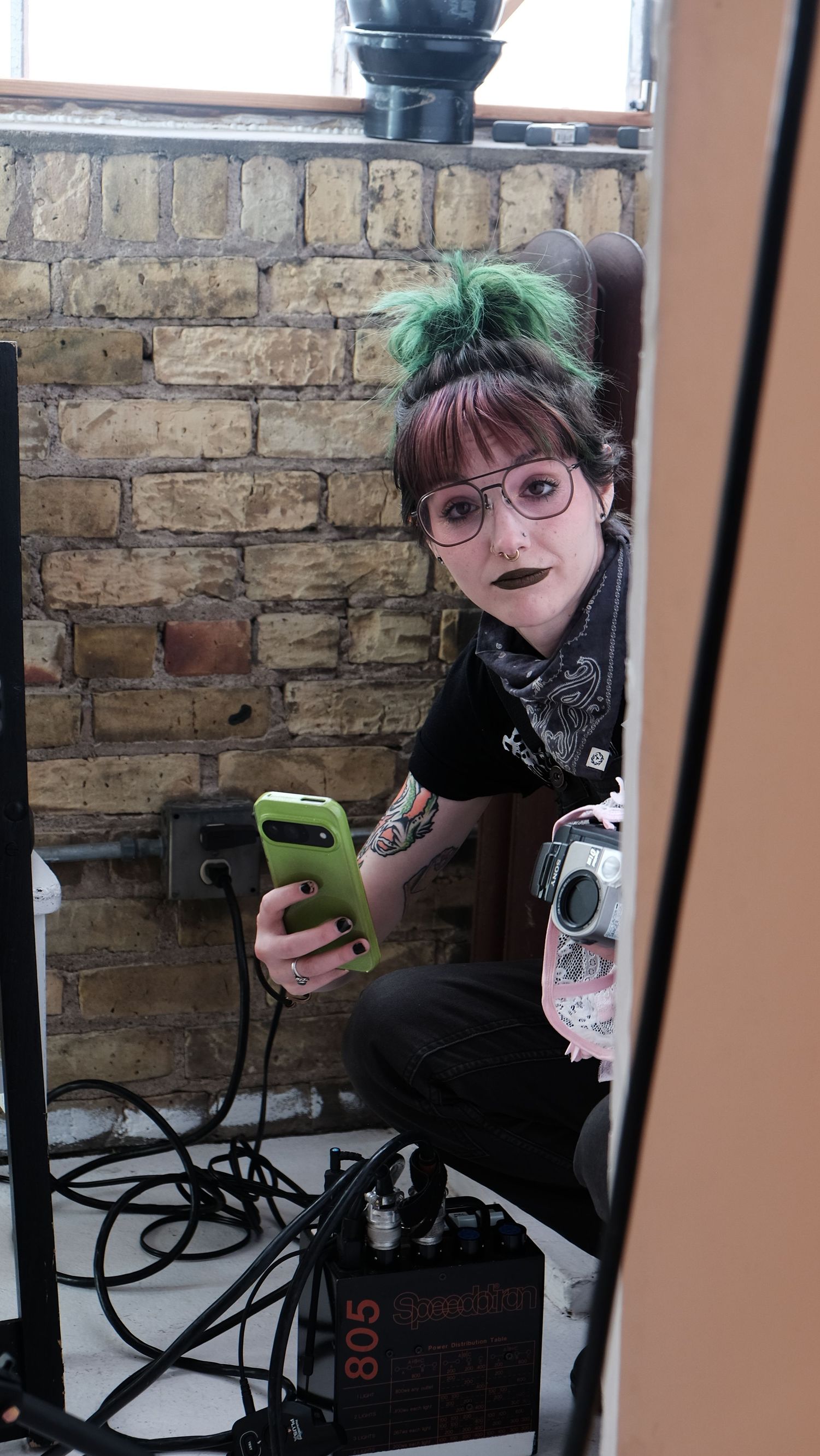 Person with green hair and glasses sits at a table looking at their phone in a dimly lit room with exposed brick walls.