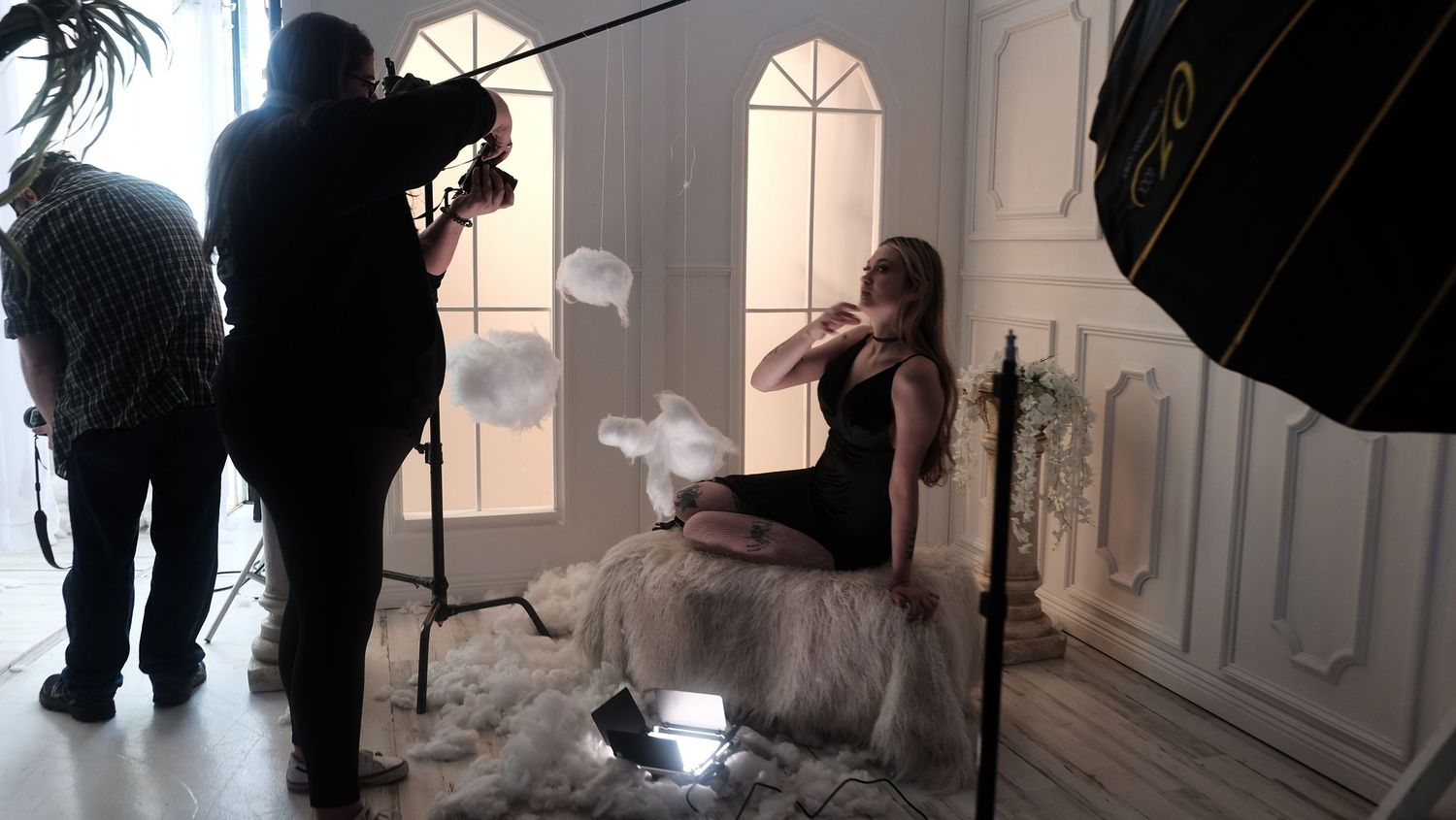 Behind-the-scenes film crew members work with lighting and camera equipment in an elegant white room.