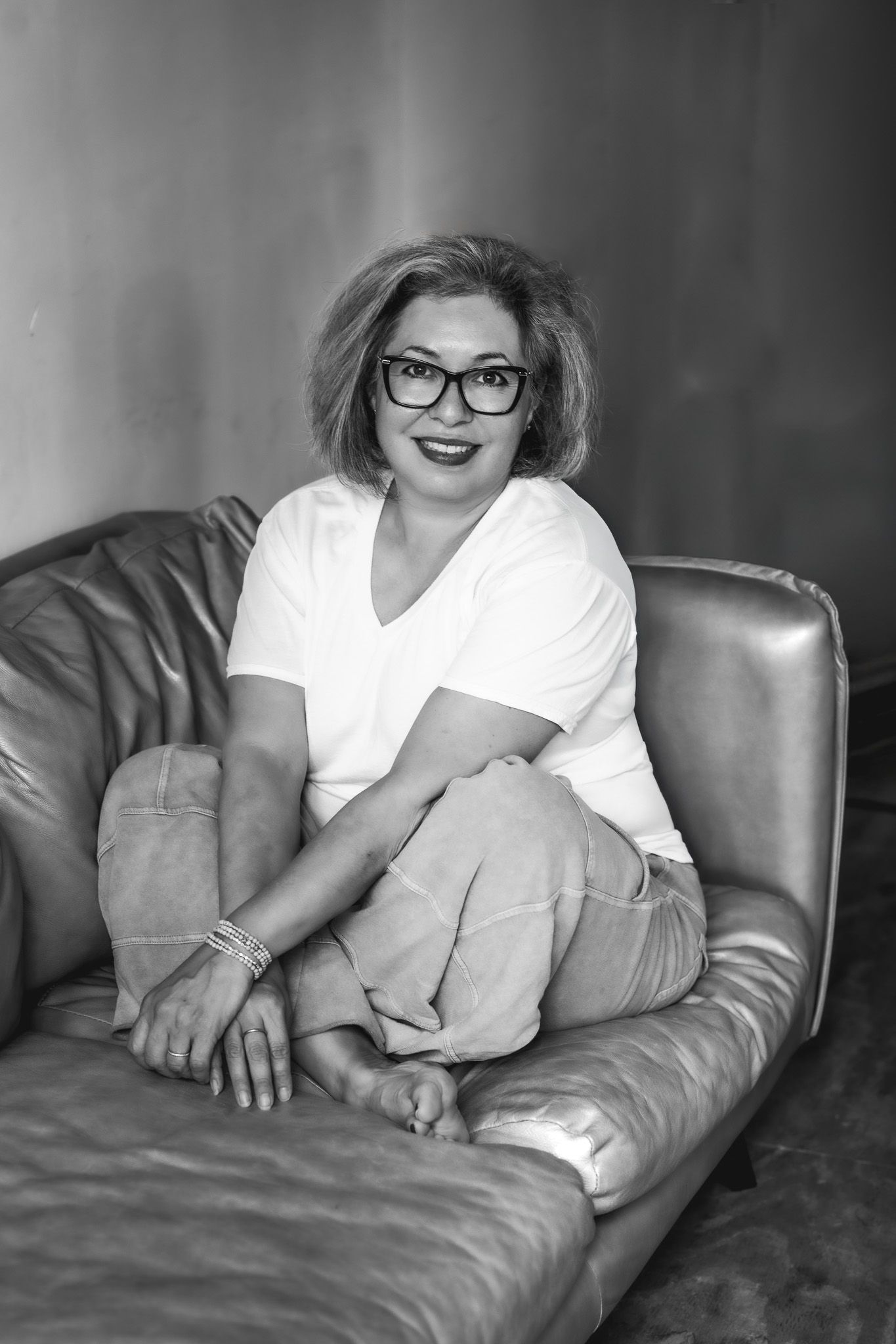 Black and white portrait of a smiling woman in glasses sitting cross-legged on a leather sofa.