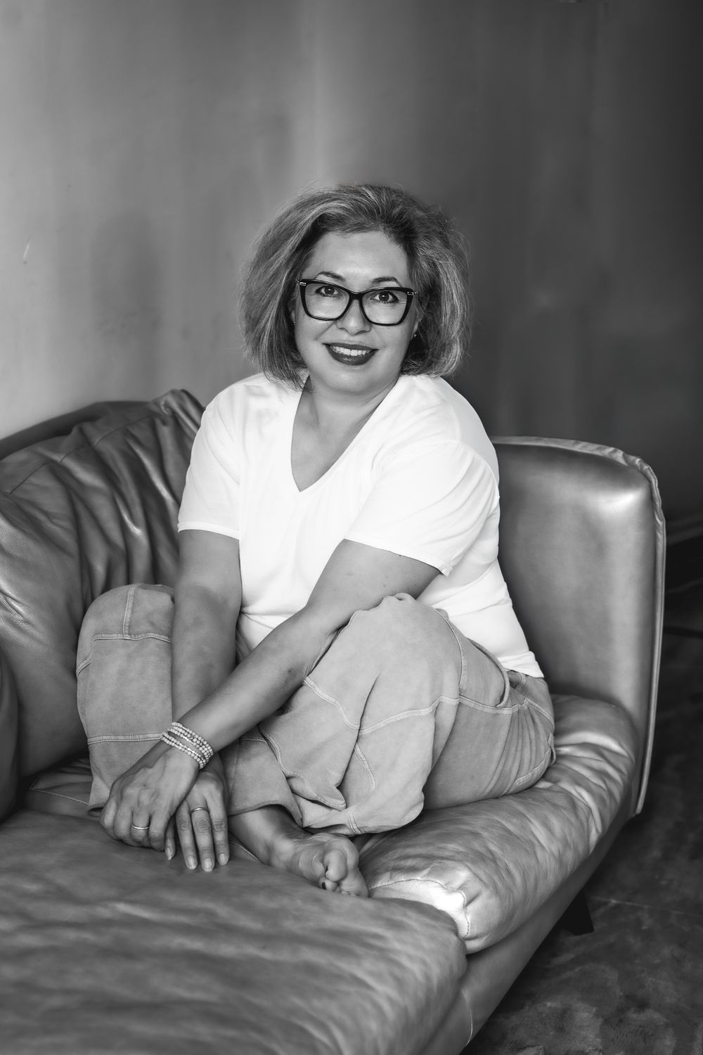 Black and white portrait of a smiling woman in glasses sitting cross-legged on a leather sofa.