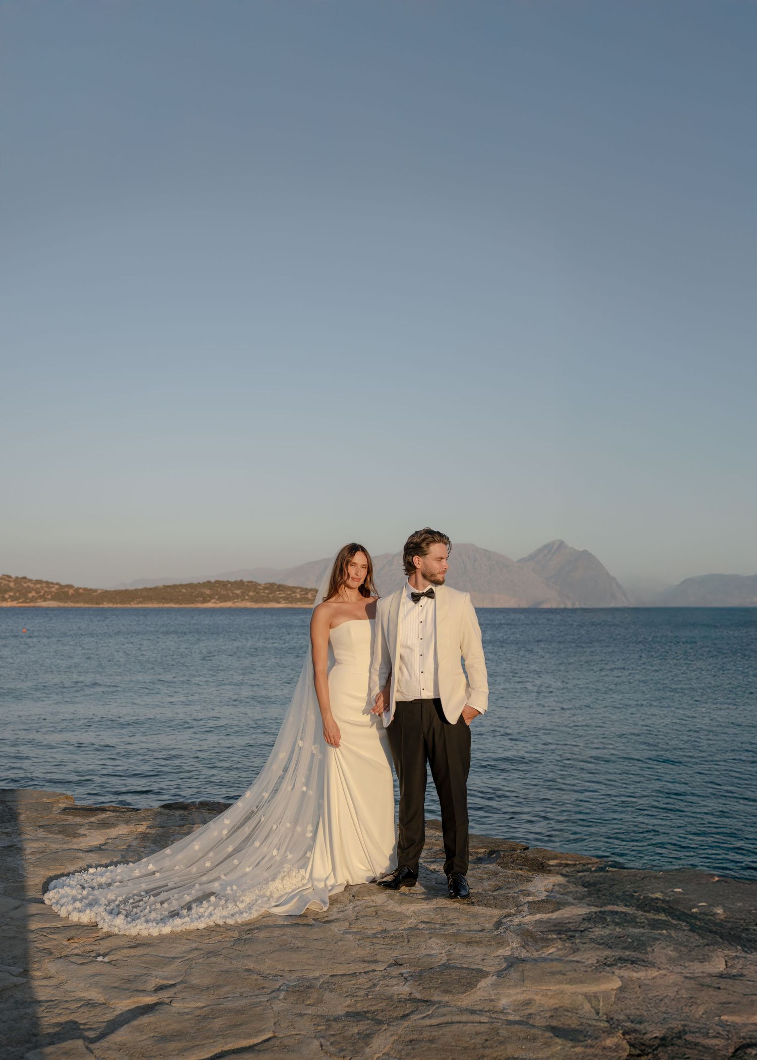 A couple in wedding attire stands on rocky coastal cliffs overlooking the Mediterranean Sea at sunset.