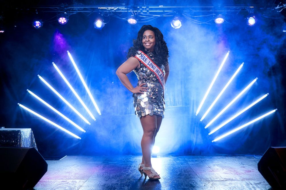 Performer in sparkly dress strikes pose on stage with blue lighting and dramatic light beams.