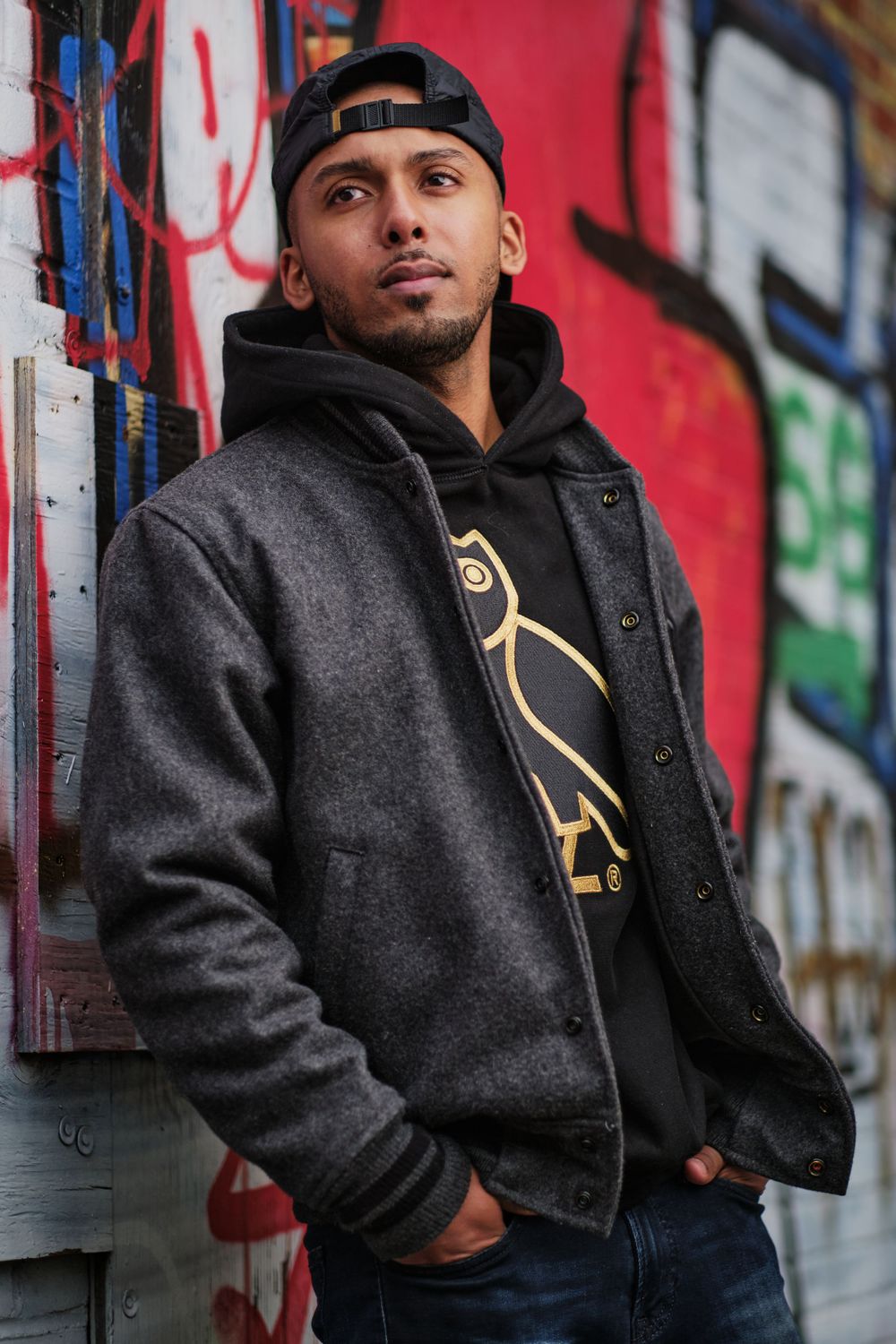Urban portrait against colorful graffiti wall featuring black hoodie and backwards cap.