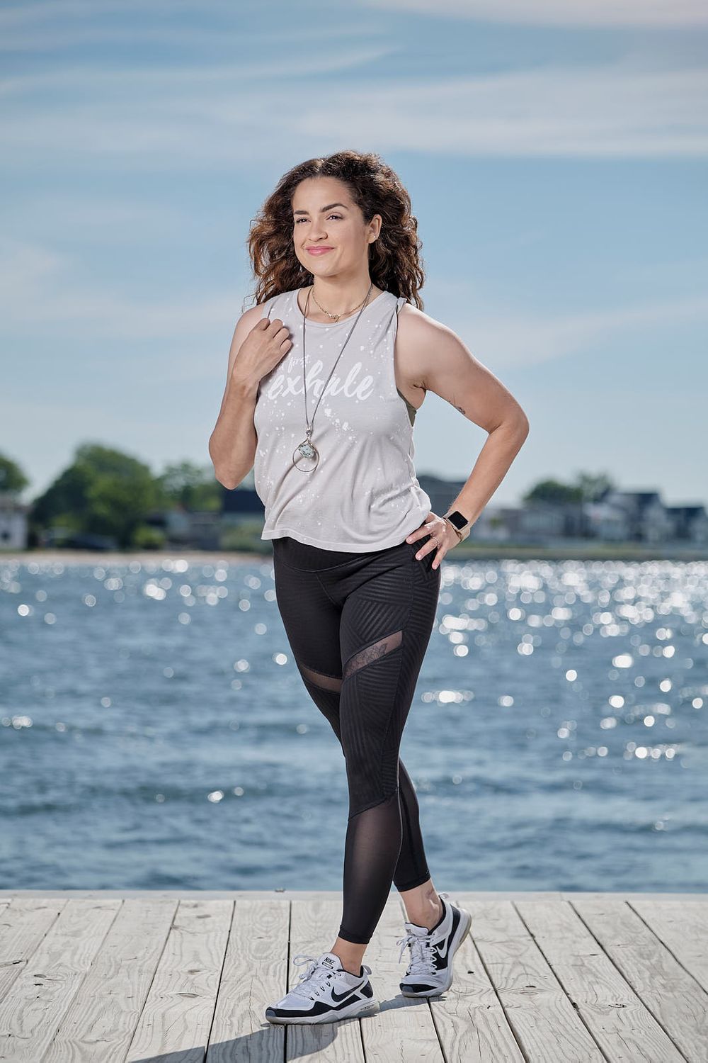 Athletic woman in workout attire poses on wooden dock with sparkling water in background.