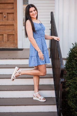 A High School Senior in a blue floral dress and white platform sandals poses on white steps with a railing.