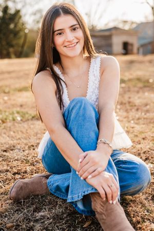 A High School Senior in a white lace top and jeans sits casually on the ground at sunset wearing brown boots.