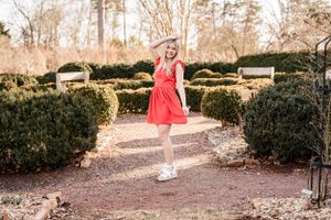 A High School Senior in a bright coral dress stands on a garden path surrounded by manicured hedges on a sunny day.