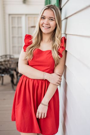 A High School Senior wearing a red summer dress with ruffled cap sleeves stands against a white wall on a porch.