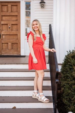 A High School Senior in a bright red summer dress and white sandals stands on white steps with a dark wooden handrail.