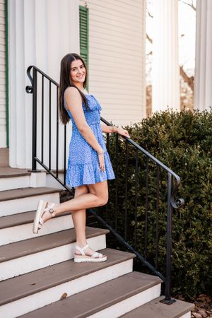 A High School Senior poses on outdoor steps wearing a blue floral sundress and white platform sandals.