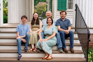 Family of five smiling on porch steps of a historic home, dressed in casual attire for an outdoor portrait.