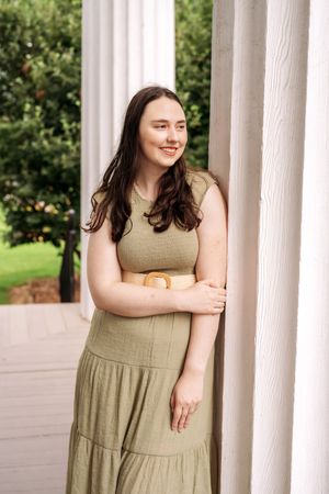 Young woman in olive green dress leaning against white column on a porch, smiling softly.