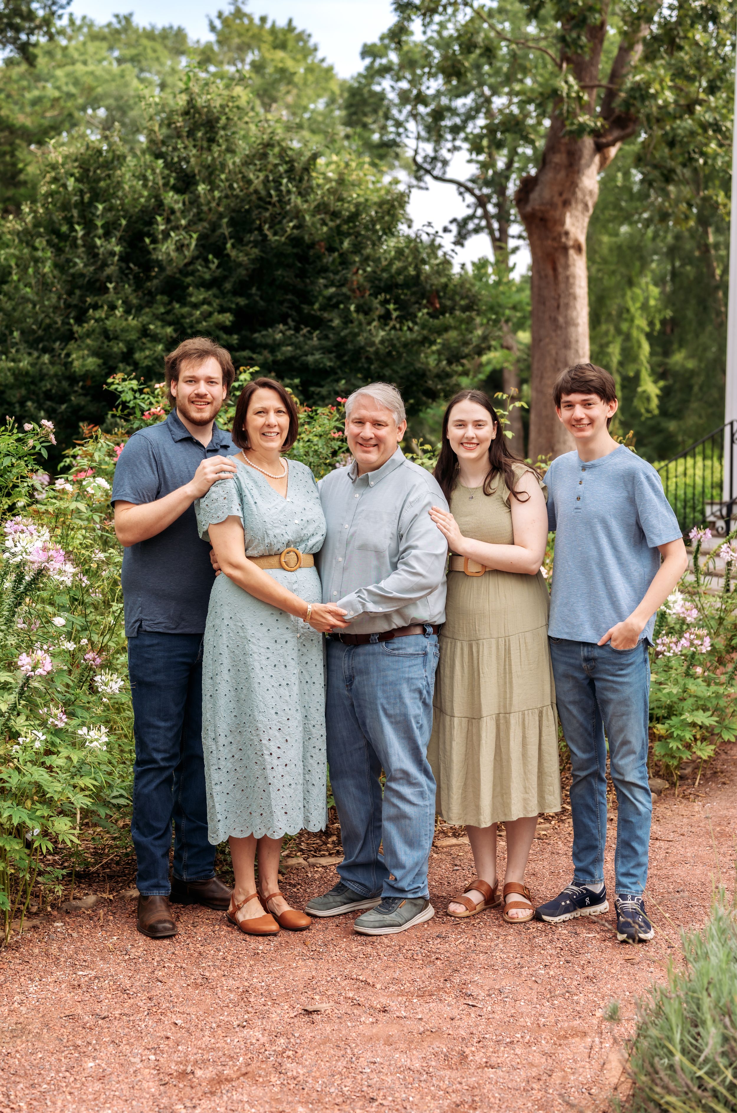 Family of five smiling together in a garden with pink flowers and lush green trees in the background.