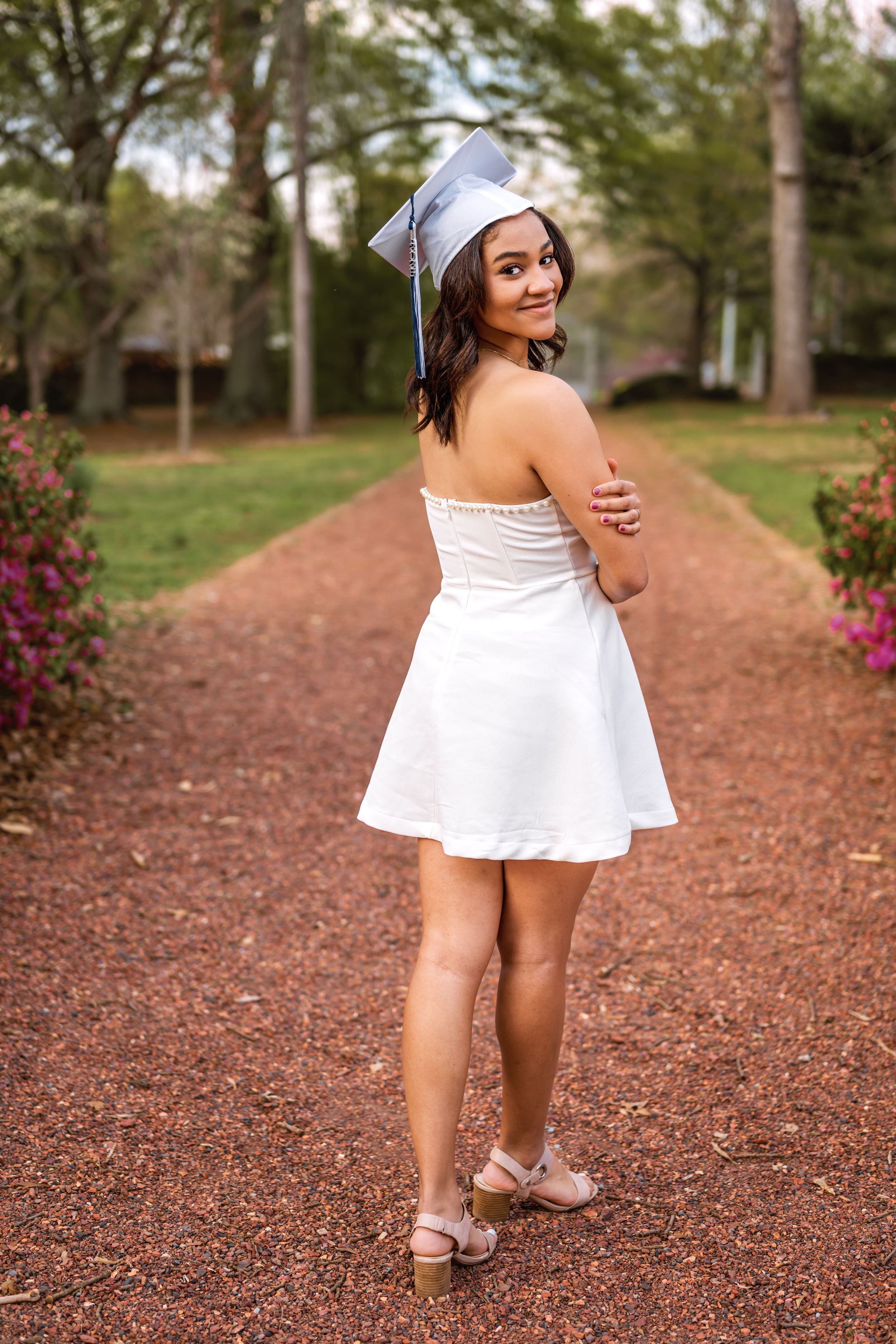 Young woman in white dress and graduation cap poses on a tree-lined path, smiling over her shoulder.