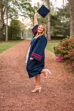 Smiling graduate in navy blue cap and gown raising mortarboard on a tree-lined path during outdoor graduation photo session.