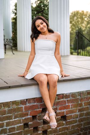 Young woman in white strapless mini dress and heels sitting on brick ledge by white columns outdoors.