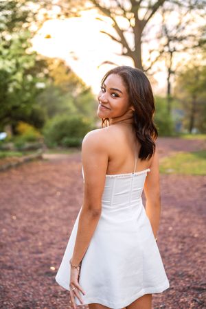 Young woman in white strapless mini dress poses outdoors at golden hour, glancing back over shoulder with a smile.