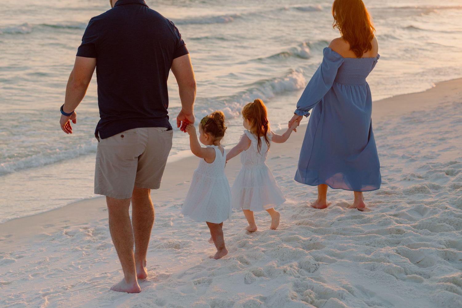 Family holding hands while walking along ocean shore at sunset.