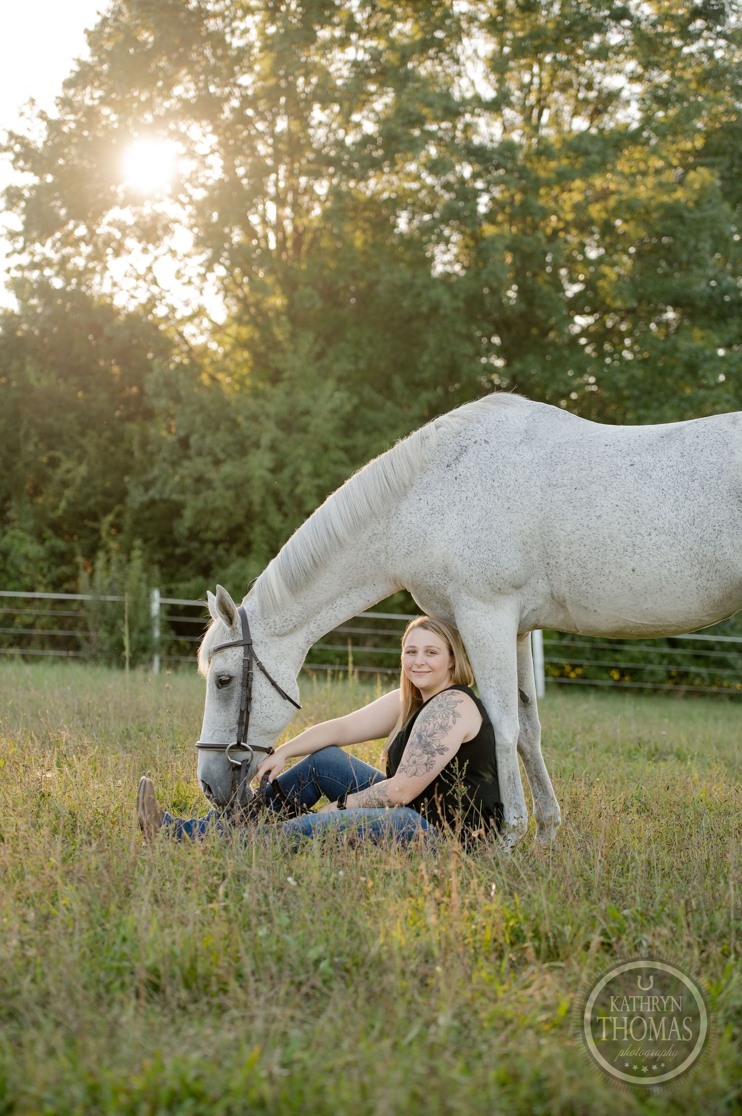 A person relaxes in the grass next to a white horse in a fenced pasture.