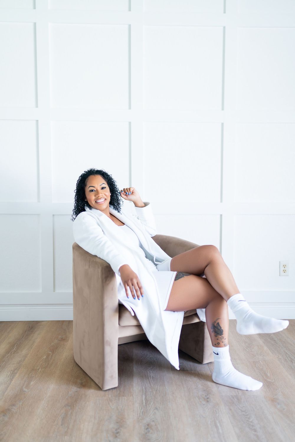 Relaxed figure lounging in beige chair wearing white robe and socks against minimalist white wall and wood floor.