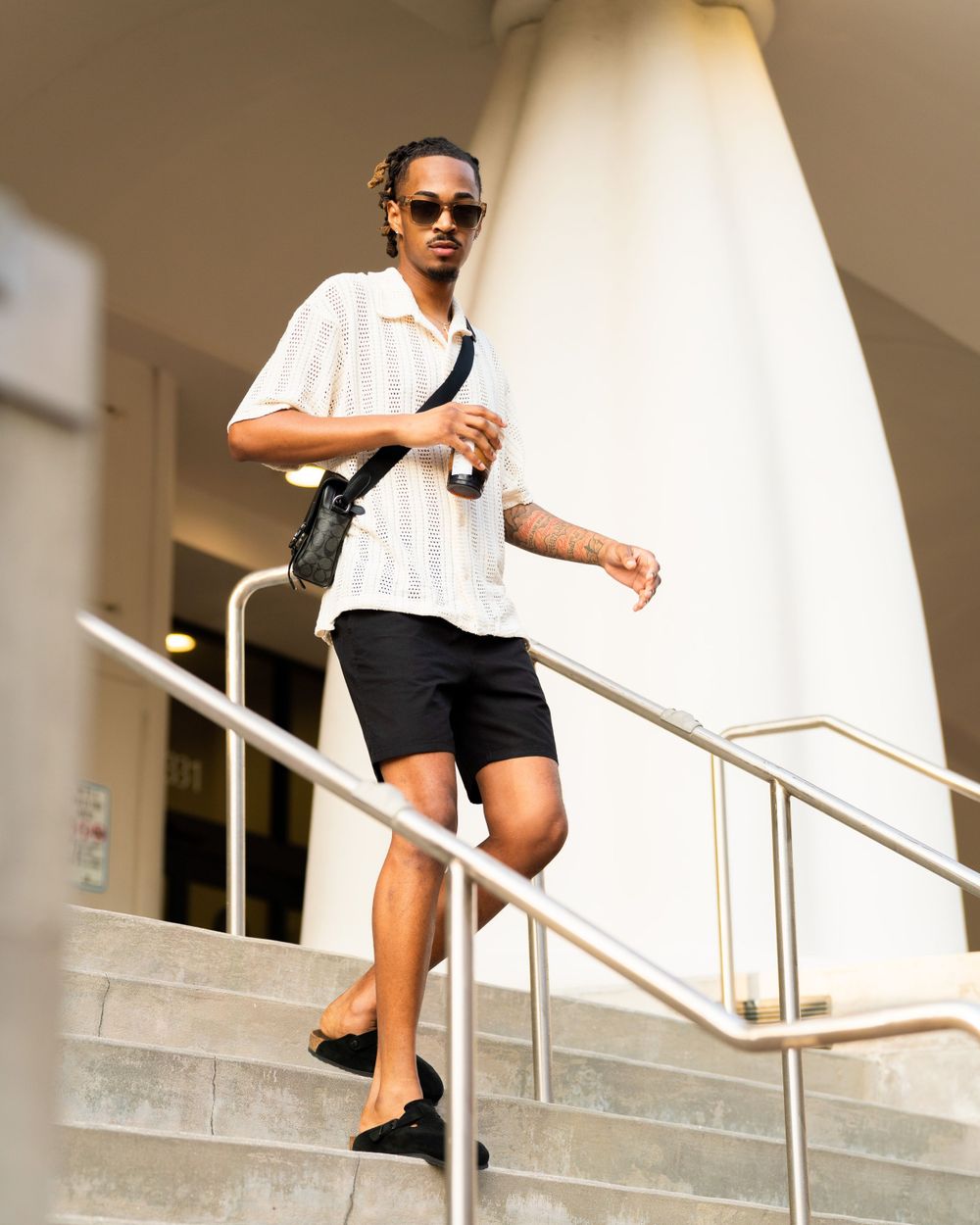 Person in a casual white shirt and black shorts walking down stairs while wearing sunglasses and carrying a bag.
