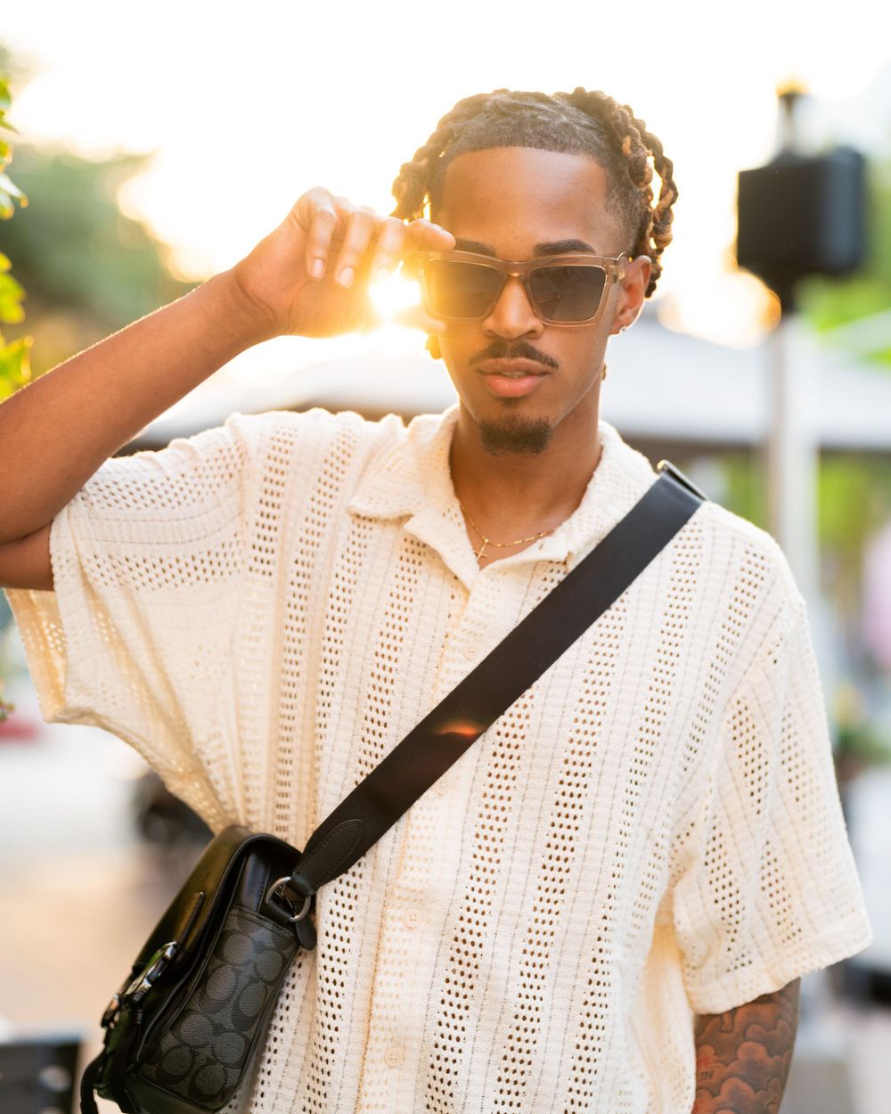 Person in cream knit sweater and sunglasses poses outdoors at sunset with crossbody bag.