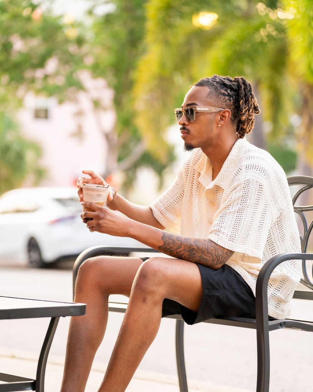 A person in a white knit sweater and shorts sits on a metal chair outdoors while holding a drink.