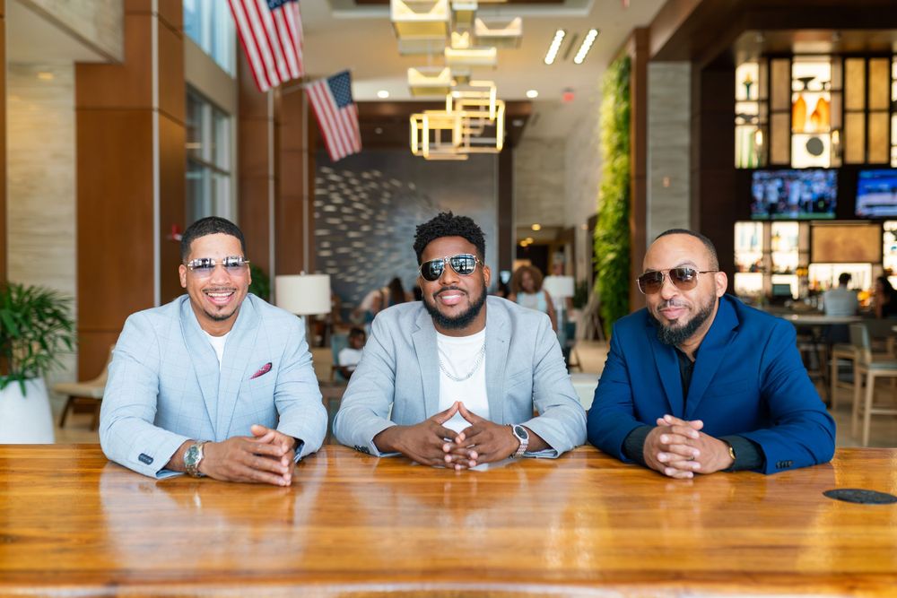 Three business professionals in casual attire sit together at a wooden table in a modern office space with American flags.