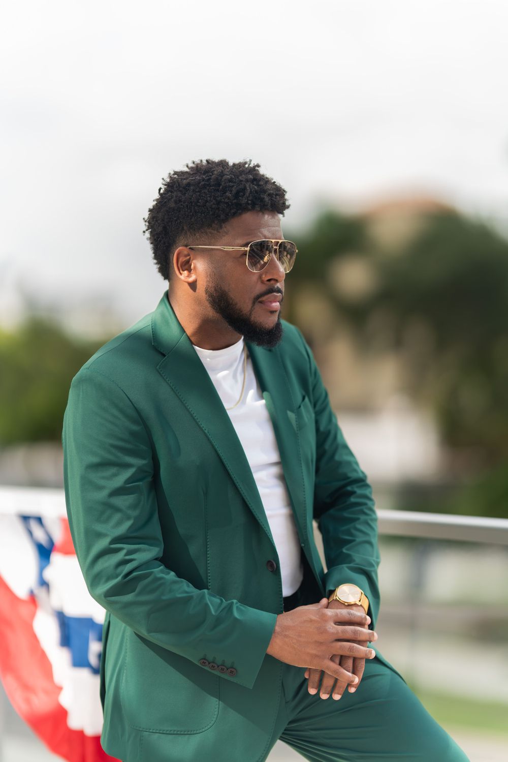 Stylish man in emerald green blazer and white t-shirt poses outdoors wearing aviator sunglasses.