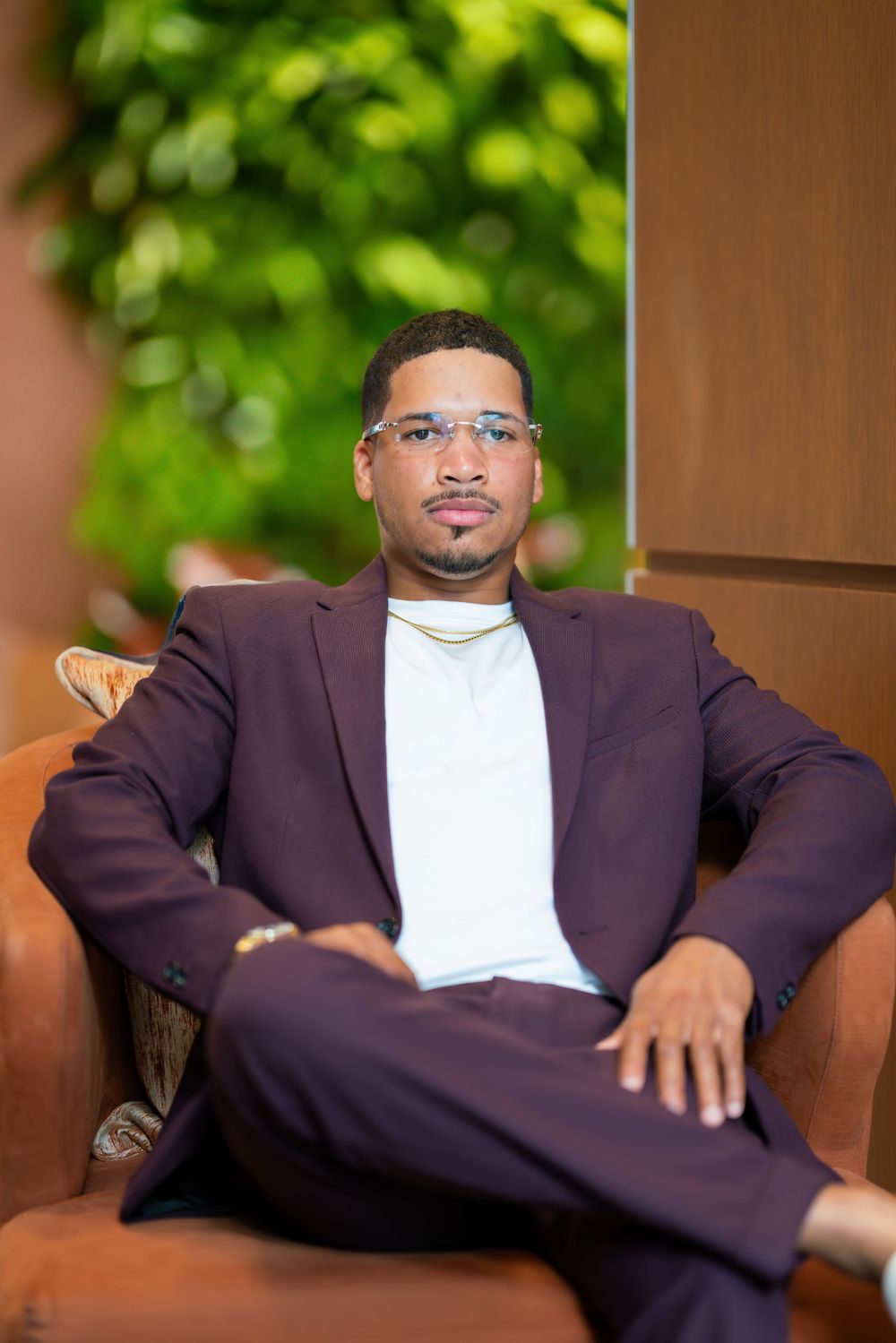 Professional portrait of someone in a burgundy suit and glasses sitting casually in a brown leather chair outdoors.