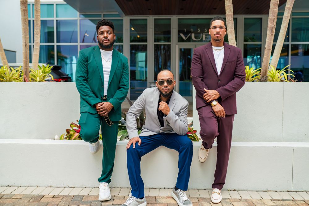 Three stylishly dressed men in colorful suits pose on outdoor steps in front of a building with windows.