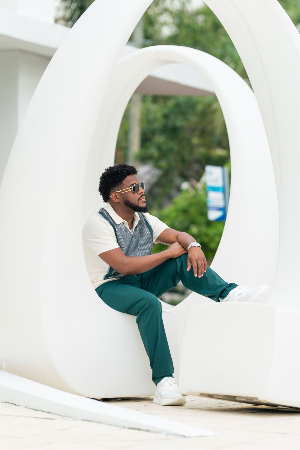 A person in sunglasses wearing a gray vest and green pants sits casually in a curved white architectural structure.