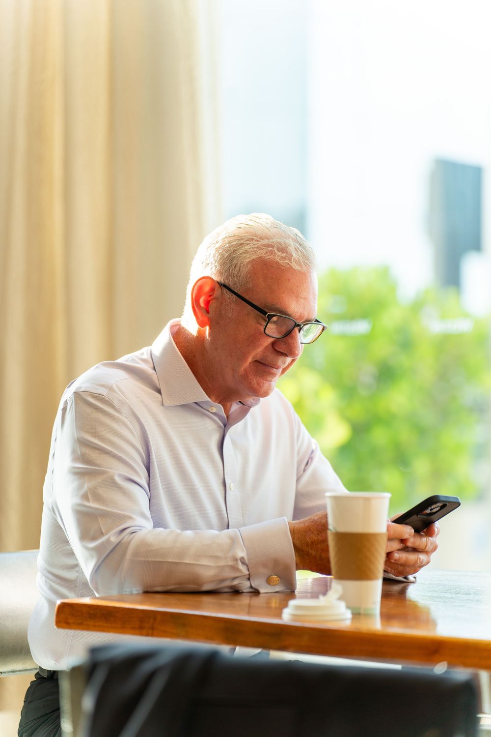 Senior businessman in glasses checks smartphone while enjoying coffee at cafe table near window with city view.