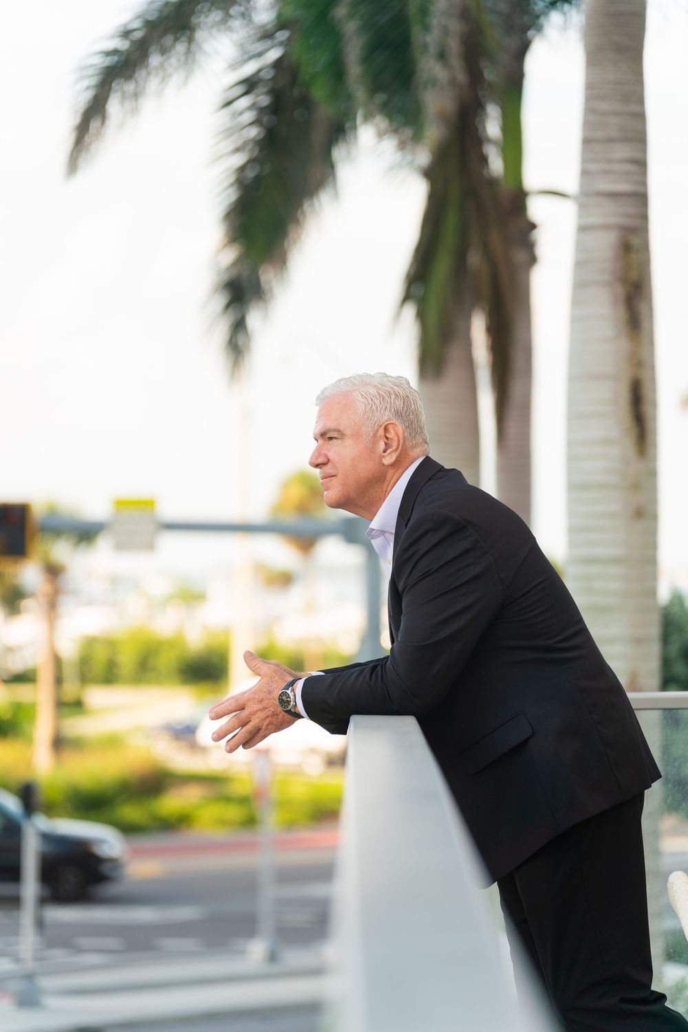 A person in a dark suit leans on a railing with palm trees and a parking lot in the background.