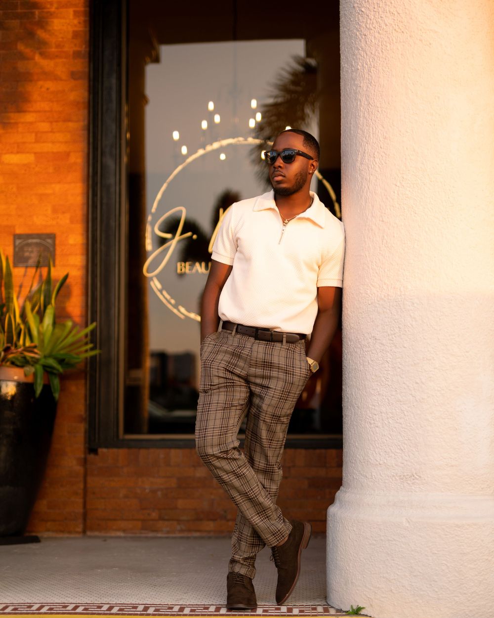 Stylish man in white polo and plaid pants poses confidently outside storefront with glowing logo and ambient lighting.