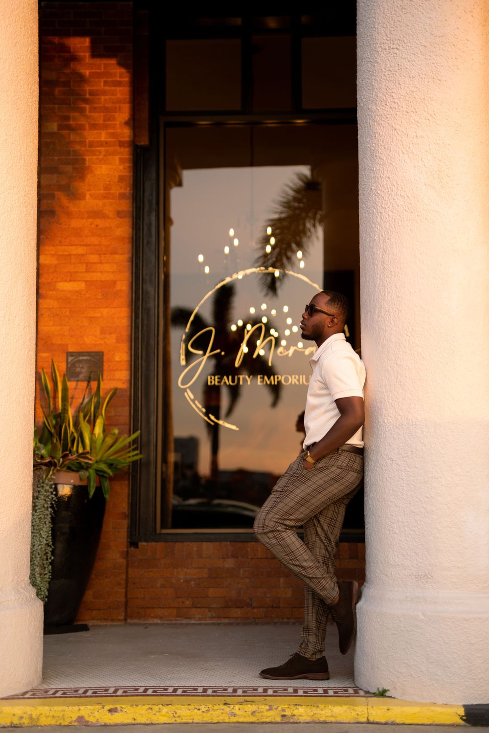 A person in a white shirt and striped pants leans against a doorway of a beauty salon with gold lettering on the window.