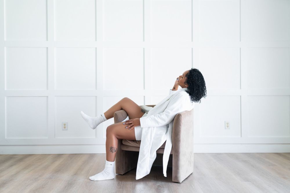 Minimalist interior scene with someone resting in a beige armchair wearing a white robe and sneakers.