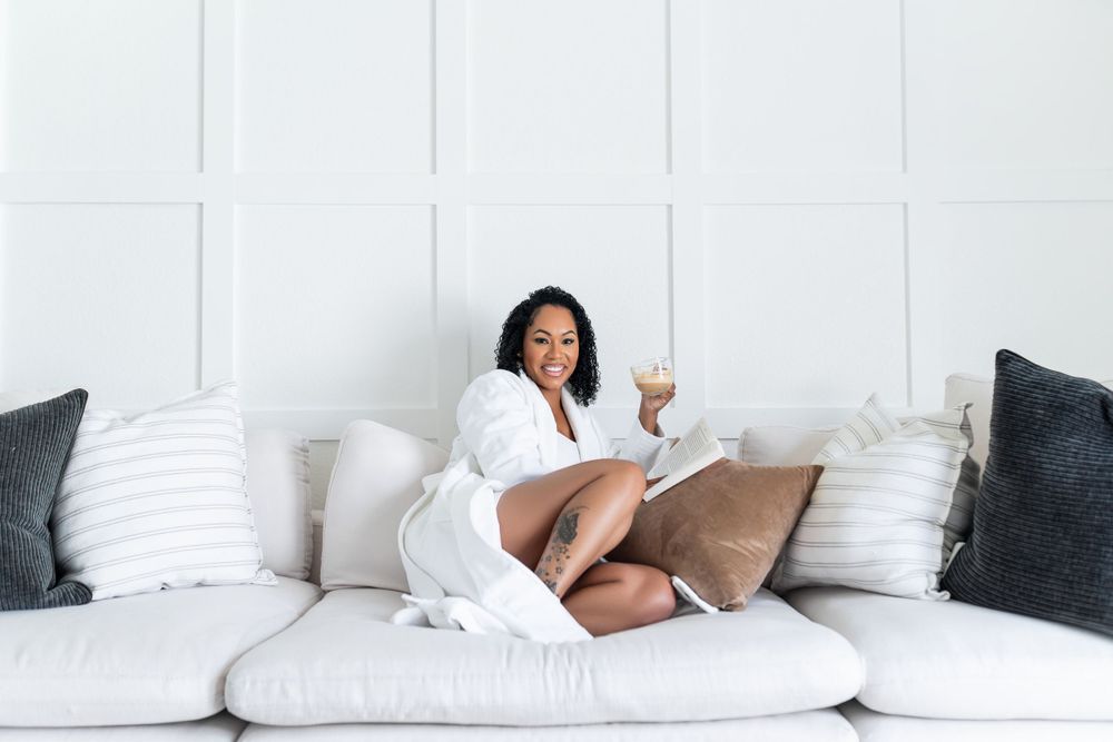 A person relaxes on a white couch with throw pillows while reading a book in a bright, minimalist living room.