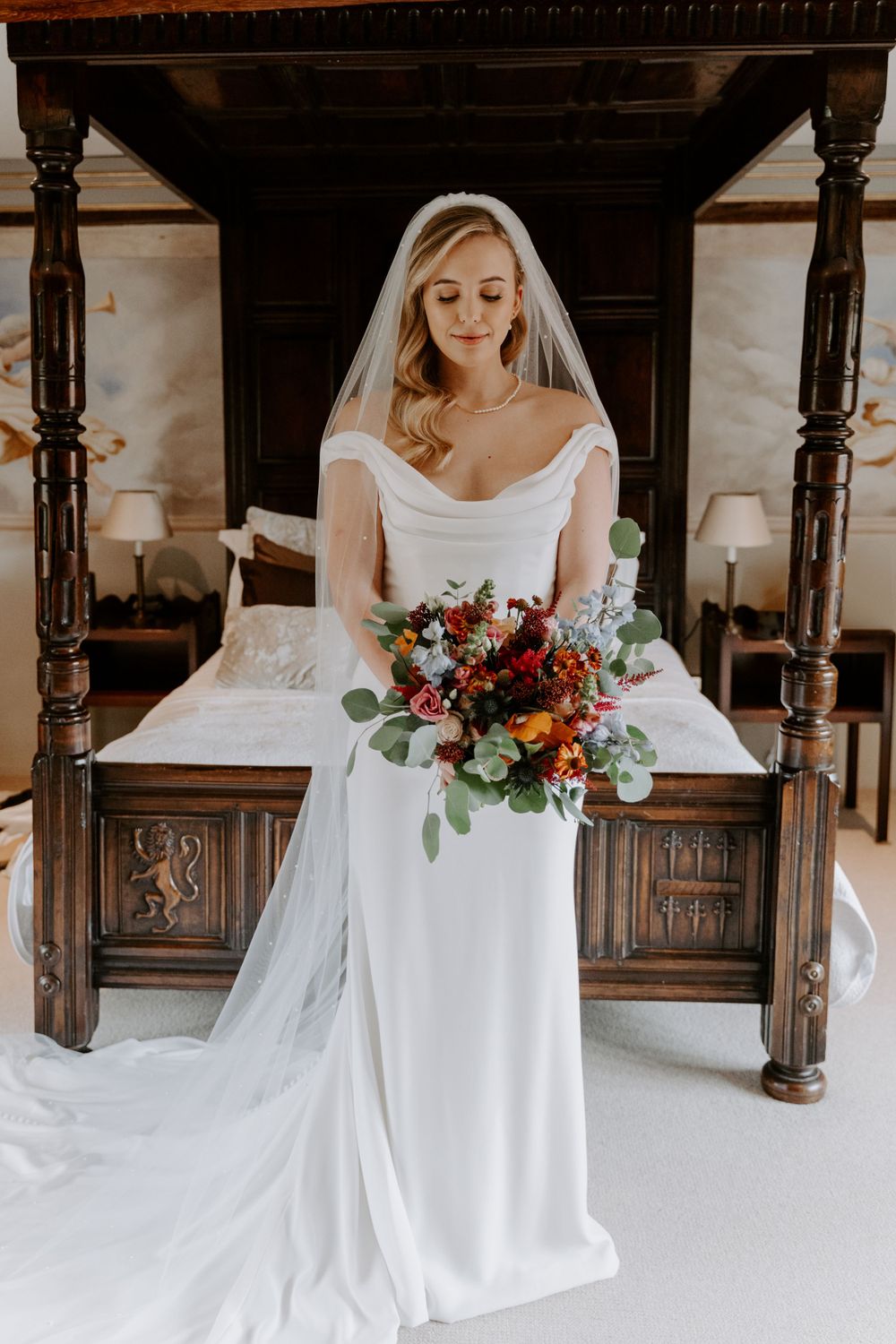 A bride in a figure hugging ivory gown is holding  her autumnal bouquet and looking down at it. Her blonde hair is in Hollywood waves.