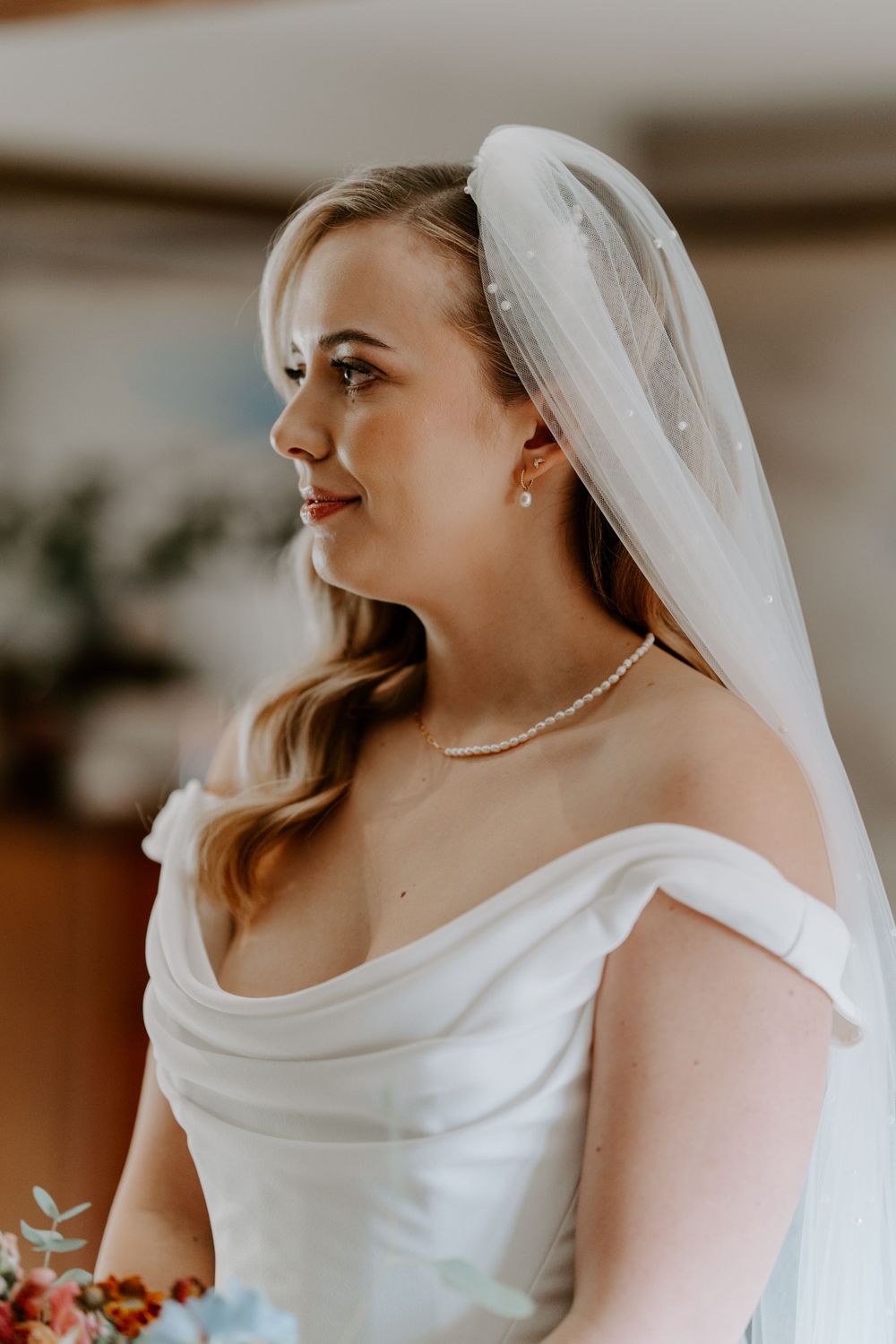 A profile view of a bride. Her blonde hair is in Hollywood waves and she has a veil in her hair. She is wearing a pearl choker necklace.