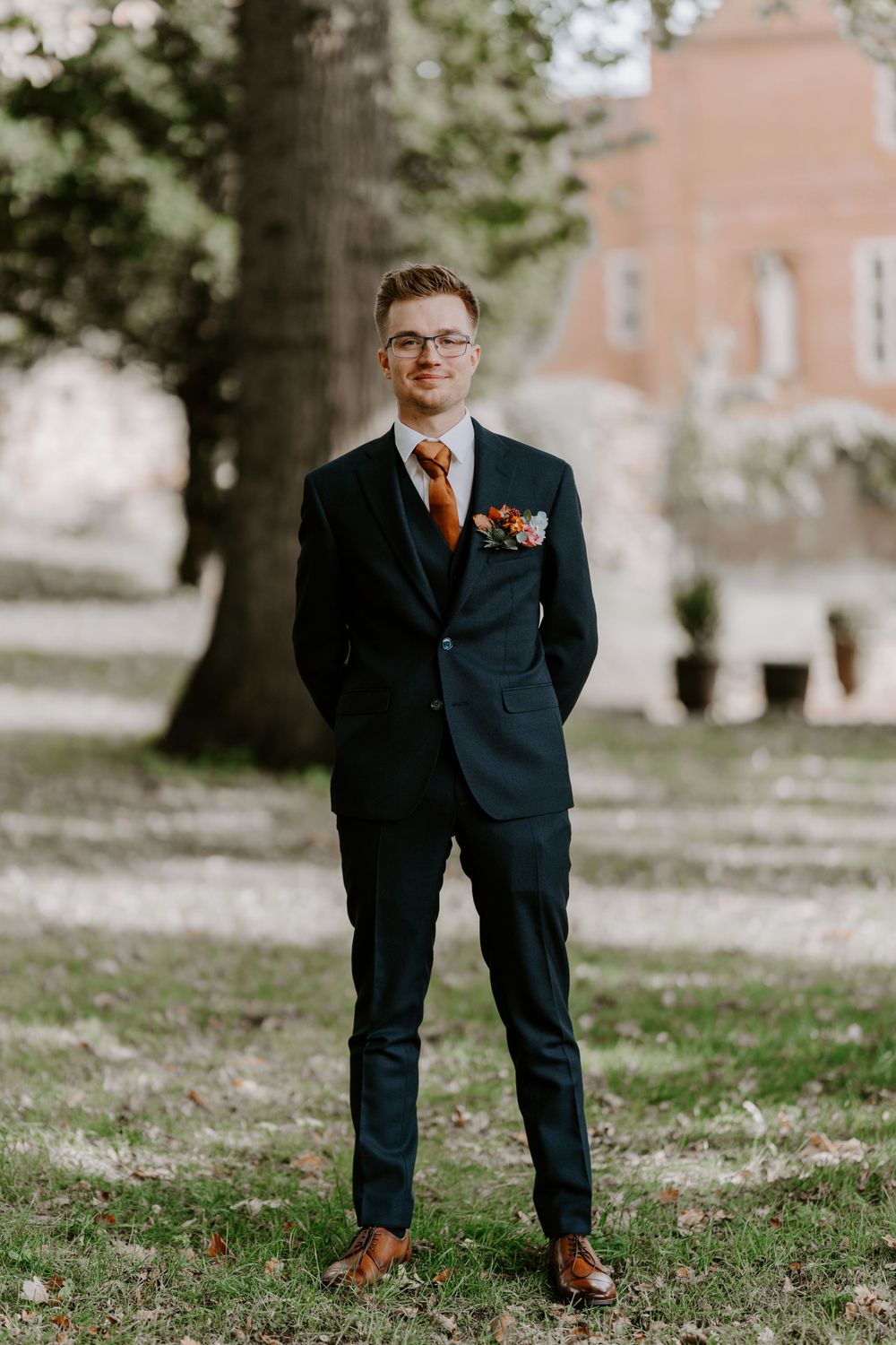 A full length photo of a groom in a navy suit and burnt orange tie. He is standing in the gardens of oxnead hall in Norfolk.