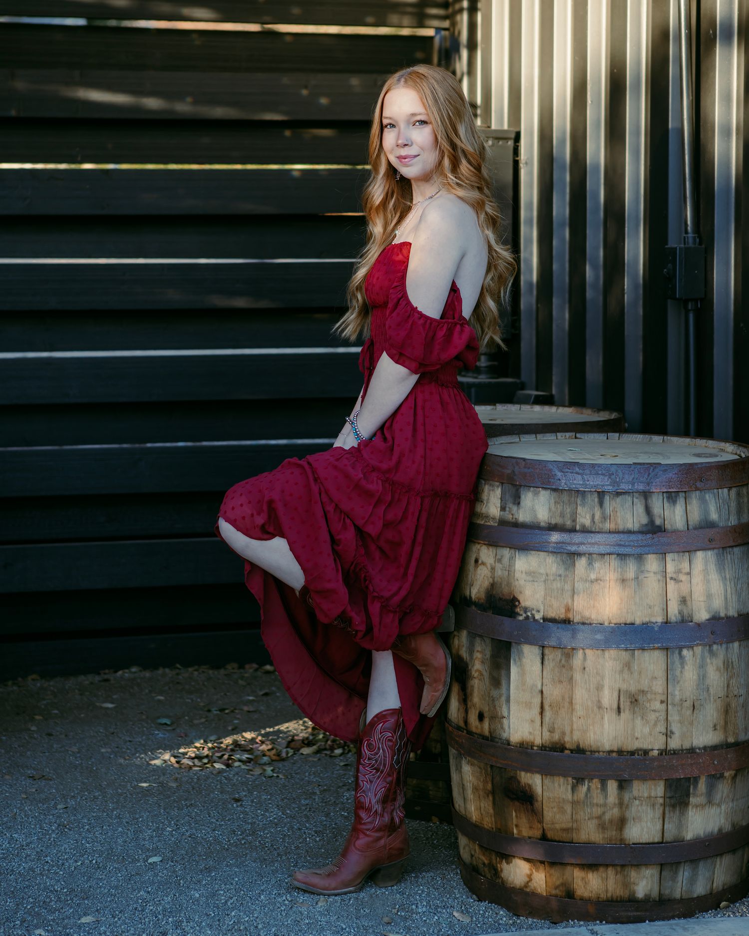 A person in a burgundy dress poses playfully near wooden barrels against a dark wall.