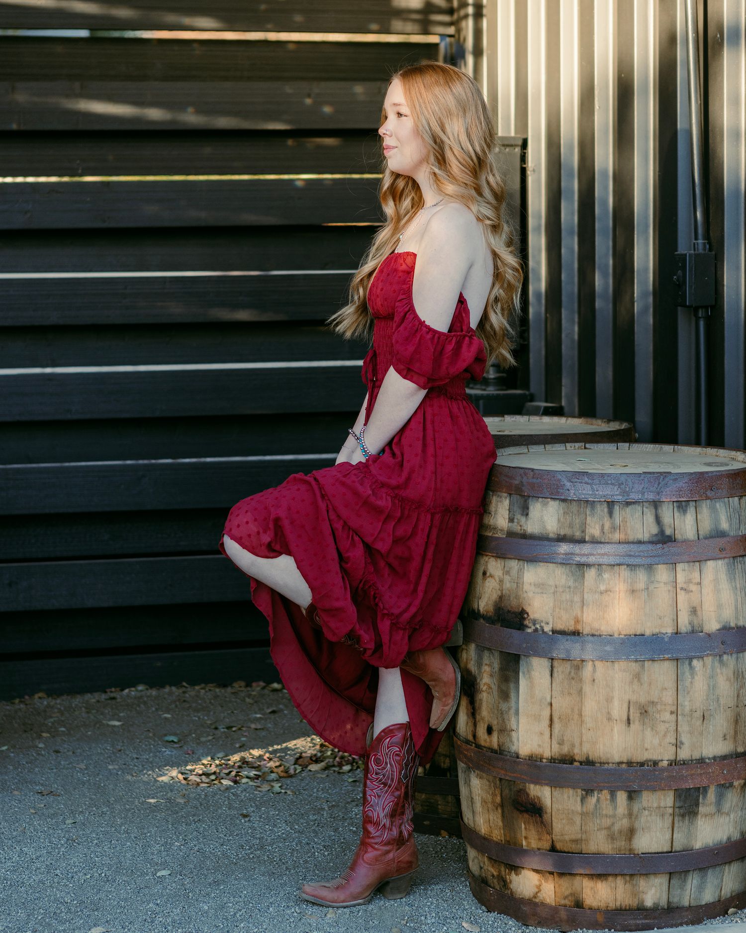 Someone in a flowing red dress and boots sits casually on a rustic wooden barrel.