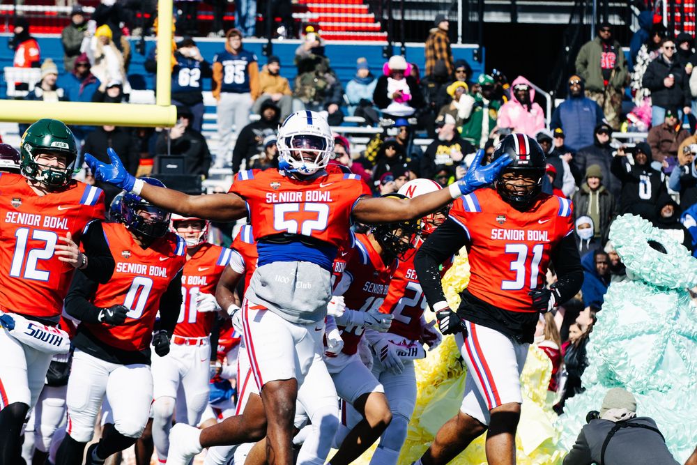 Senior Bowl football players in red uniforms celebrate on the field during a sunny college all-star game.