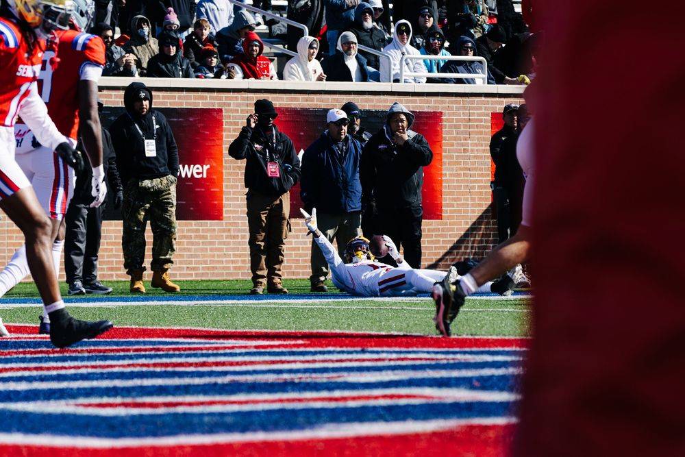 Football player diving near the sideline during a college football game with spectators and photographers watching.