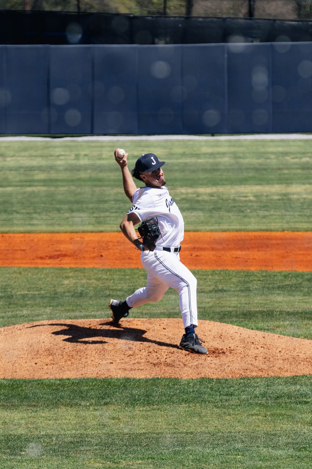 Baseball pitcher in white uniform follows through on delivery motion during game on sunny field.