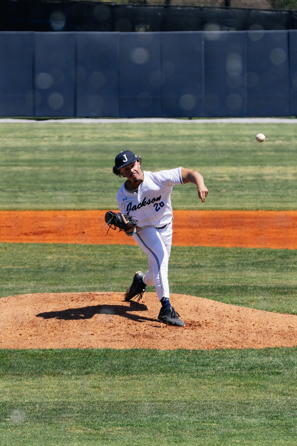 Baseball pitcher in white uniform follows through on delivery from the mound during a sunny game on grass and clay field.