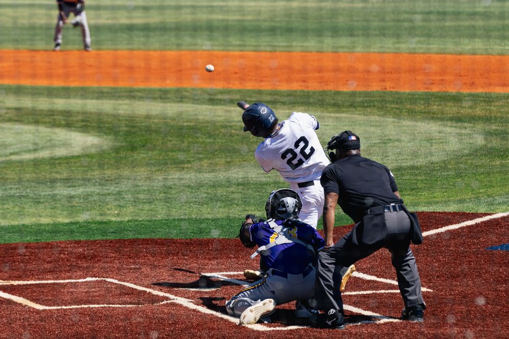 Baseball batter at home plate swings at a pitch during a game on a sunny day with green outfield in background.