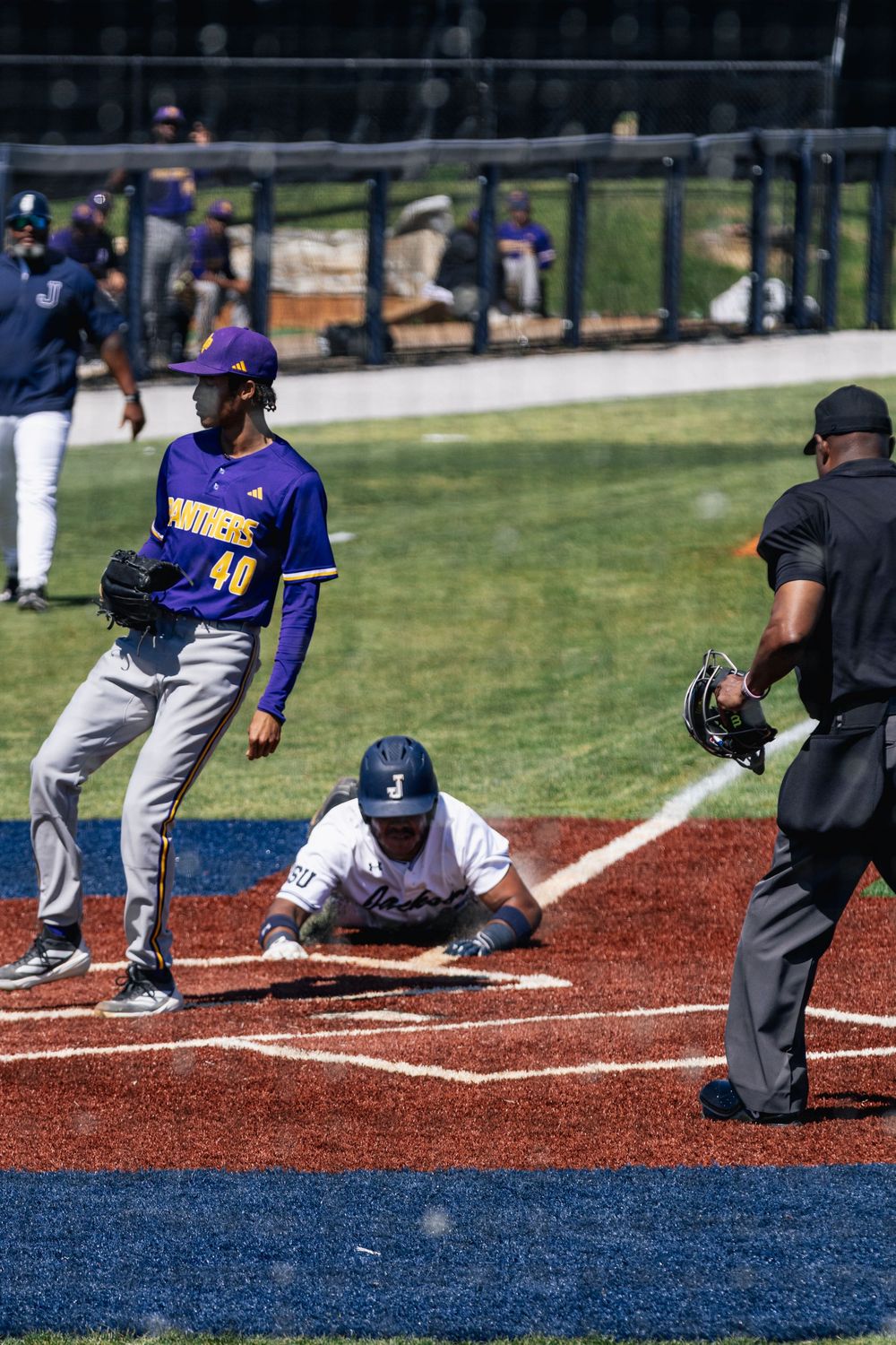Baseball player in purple uniform slides into base during game while umpire watches from behind home plate.