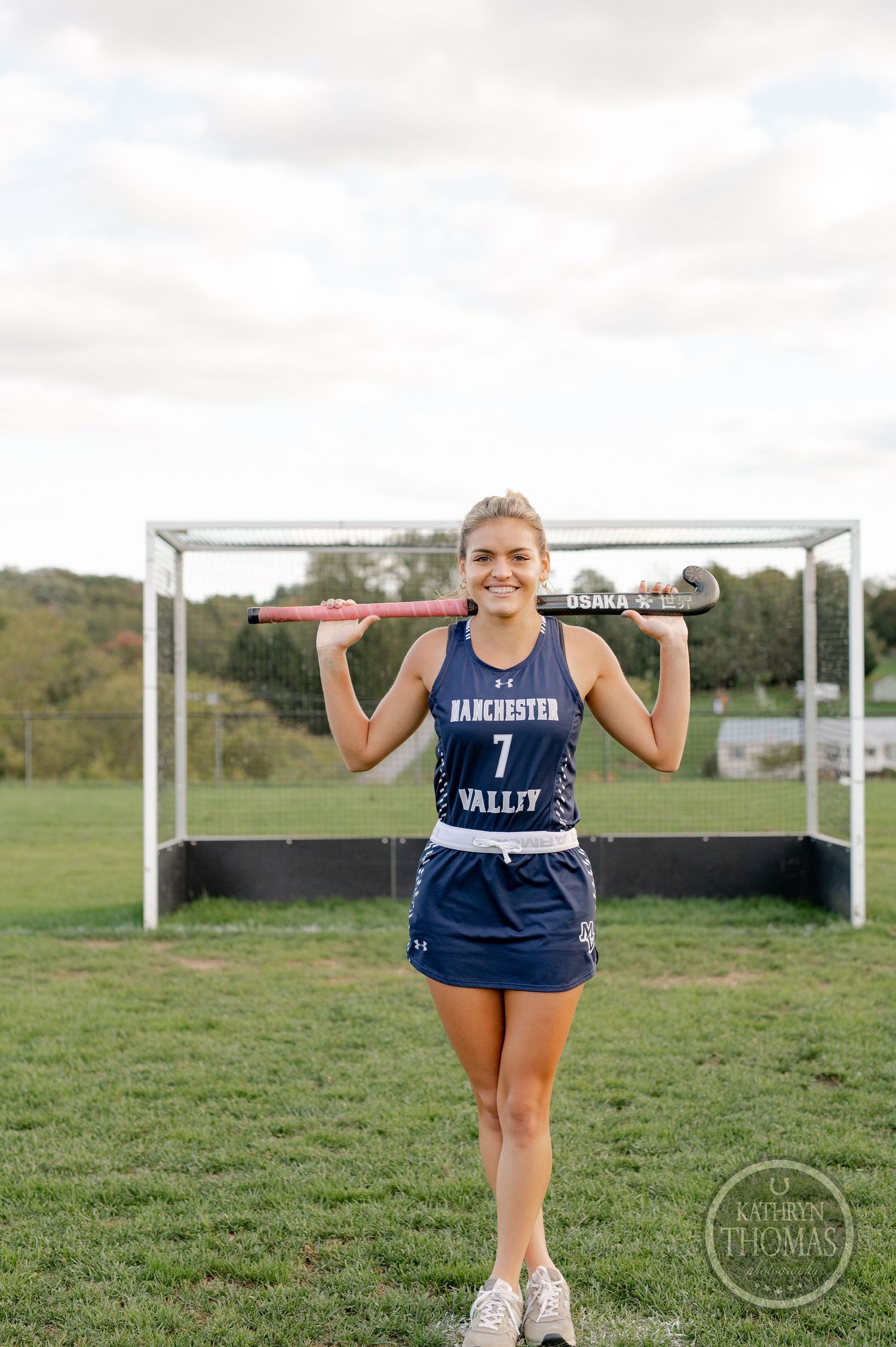 Field hockey player in blue uniform poses with stick in front of goal on grass field.